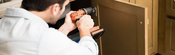 Een persoon die met een oranje accuboormachine een bruine houten kast onder een aanrecht in de keuken repareert of in elkaar zet, heeft een geconcentreerde uitdrukking op zijn gezicht.