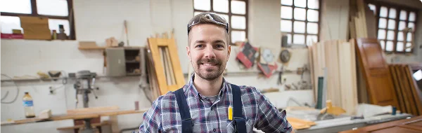 Een lachende timmerman in een werkplaats, gekleed in een geruit overhemd en gereedschapsriem, met een hamer in zijn hand. Omringd door hout en gereedschap, straalt hij zelfvertrouwen uit.