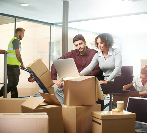 Kantoormedewerkers, omringd door dozen en computers, pakken spullen uit in een lichte kantoorruimte. Op de achtergrond is een verhuizer met een steekwagentje te zien. Een drukke, maar ook collaboratieve sfeer.